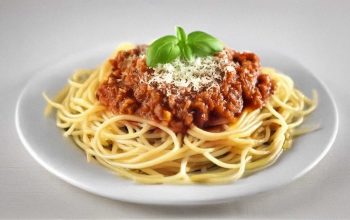 A close-up, wide-angle shot of a bowl of spaghetti Bolognese on a rustic wooden table, with grated Parmesan cheese in a small dish nearby, a fork placed to the side, and fresh parsley leaves scattered for garnish. Warm lighting emphasizes the rich, hearty texture of the sauce.