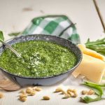 A bowl of freshly made arugula pesto on a white shabby wooden surface, with a spoon dipping into the pesto, surrounded by neatly arranged ingredients including arugula, lemon, pine nuts, Parmesan cheese, and garlic
