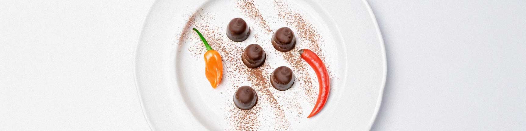 A plate of chocolate truffles dusted with cocoa powder and garnished with chili flakes and an orange habanero chili, presented on a white background.