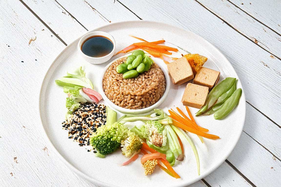 A macrobiotic dish featuring grains, colorful steamed vegetables, tofu cubes, and a small bowl of tamari sauce on a white wooden surface.