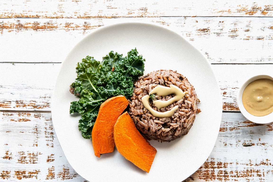 A plate with brown rice, steamed kale, roasted sweet potatoes, and tahini sauce, set on a white shabby wooden surface.