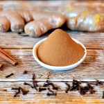 Fresh spices for pumpkin pie spice, including cinnamon sticks, nutmeg, ginger root, and cloves, arranged on a white shabby wooden surface.