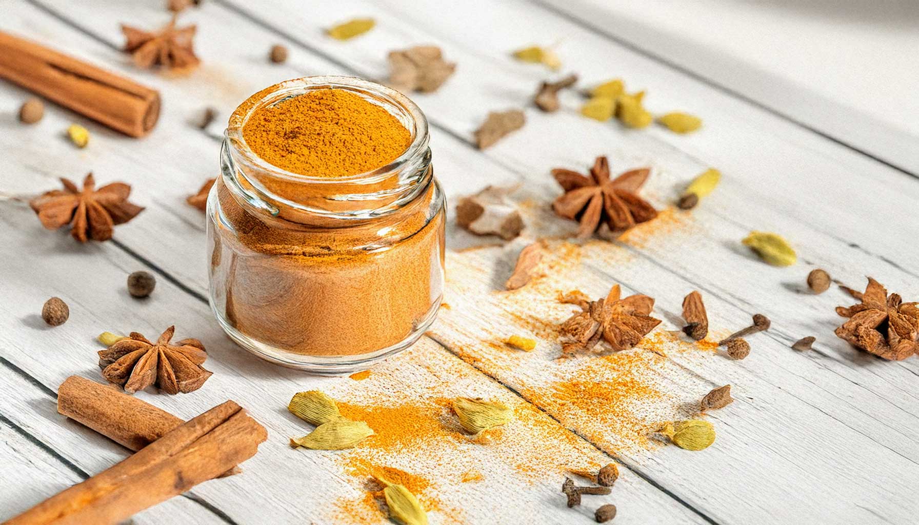 A small glass jar of homemade pumpkin spice blend, surrounded by teaspoons of individual spices on a white shabby wooden surface, illuminated by soft, natural lighting.